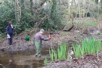 Person in waders walking through a pond and person on the bank holding rubbish collected