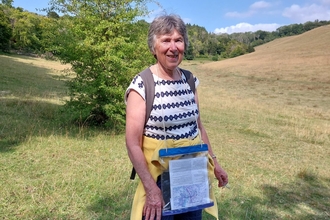 A lady walking through a grassy valley with trees in the distance