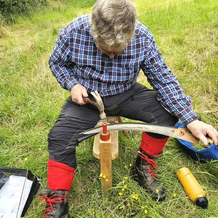 A man sat on a wooden bench peening a scythe blade.