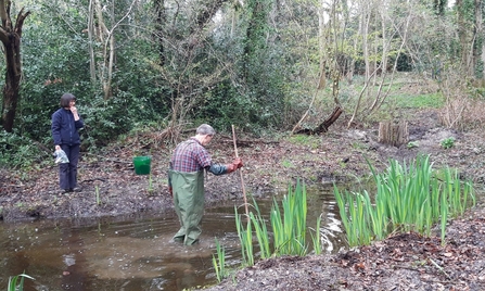 Person in waders walking through a pond and person on the bank holding rubbish collected