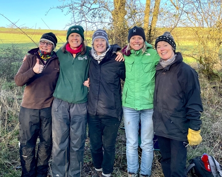 A group of five women in a woodland smiling at the camera, dressed in winter clothing